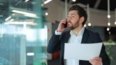 İş adamı telefonda bağırırken öfke ve hayal kırıklığını ifade ediyor. Belgeleri elinde tutmak, modern ofiste stresli ve hoşnutsuz görünmek. İletişim sorunları, çatışma ve baskı bariz.