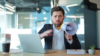 İş adamı, megafonla yüksek sesle iletişim kurarak öfke ve hayal kırıklığını ifade eder. Yoğun stres ve saldırganlık duyguları modern ofisi dolduruyor. Dikkat istiyor, acil duyuru yapıyor..