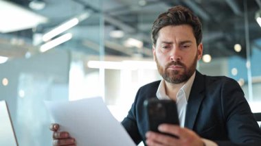 Profesyonel, çok önemli iş belgelerini akıllı telefondaki bilgilerle karşılaştırır. Ciddi davranış ve odaklanmış dikkat, ofisteki önemli verilerin derin düşünce ve analitik incelemesini gösterir.