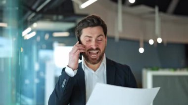 İş adamı telefonda bağırırken yoğun öfke ve hayal kırıklığını dile getiriyor, modern bir ofisteki önemli belgeleri inceliyor. İletişim sırasında çatışma ve stres duyguları açıkça görülür..