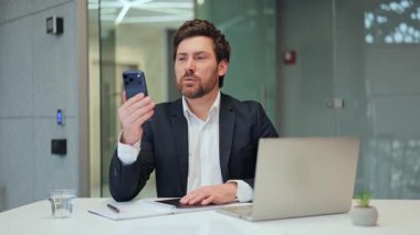 Takım elbiseli bir profesyonel akıllı telefondan görüntülü arama yapıyor. Bağlılık, güven ve mutluluğu ifade ediyor. Modern ofis arka planı sorunsuz dijital iletişimi gösterir.