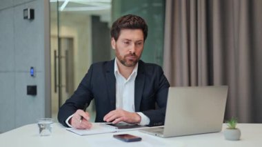 Ofis masasında özenle çalışan profesyonel bir adam. Not defteri ve dizüstü bilgisayar, devam etmekte olan iş operasyonları için yoğun çaba ve düşünceli bir değerlendirme göstererek görevin merkezindedir..