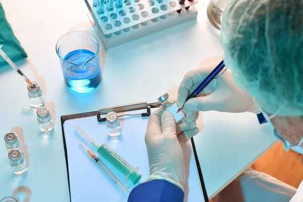 Medical worker equipped with personal protection writing on vial on laboratory table. Top view. Horizontal composition.