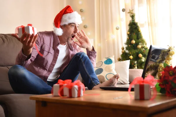 Man with gift in hand looking at laptop with surprise at christmas sitting on a sofa in the living room. Horizontal composition.