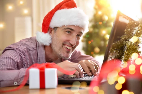 Smiling man with christmas hat using laptop at home with christmas background. Horizontal composition.