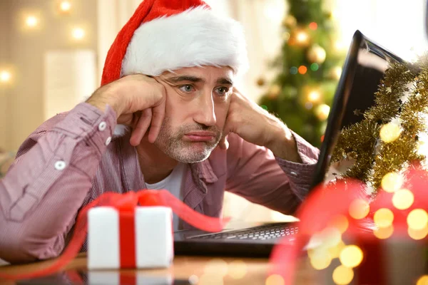 Man with christmas hat looking at laptop screen disappointed with christmas background. Horizontal composition.