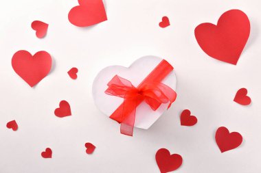 Kalp şeklinde beyaz hediye kutusu ve kırmızı fiyonk ve beyaz masada kırmızı kalplerin kesikleri. Üst görünüm.