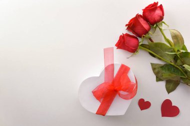 Üç kırmızı gül ve kırmızı kalp şeklinde yaylı beyaz bir hediye kutusu ve beyaz bir masada kırmızı kalplerin kesikleri olan romantik bir arka plan. Üst görünüm.