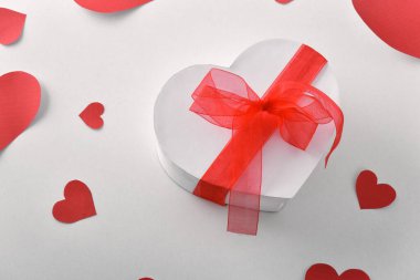 Kalp şeklinde beyaz hediye kutusu ve kırmızı fiyonk ve beyaz masada kırmızı kalplerin kesikleri. Yüksek görünüm.
