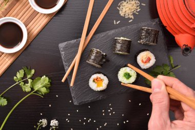 Elleri çubuklu, tahta tabaktan suşi toplayan, içinde malzemeler, kaplar ve karanlık arka plan olan sofralar. Üst Manzara. Yatay bileşim.