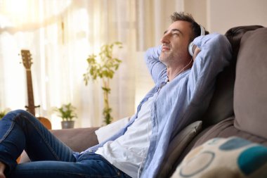 Rahatlamış bir adam, beyaz kulaklıklardan müzik dinlerken evdeki kanepede boş boş oturuyor.