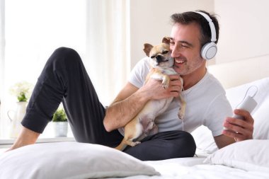 Chihuahua köpeğini okşayan bir adam yatağın üzerinde duran kulaklıklı bir cep telefonundan müzik dinliyor.