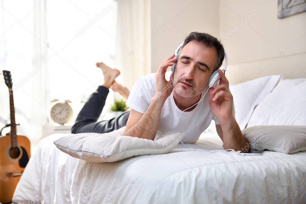 Music lover listened concentrated lying on the bed from a mobile with white headphones with guitar and window in the background entering the sunlight