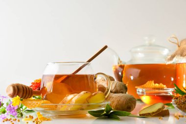 Zencefil kökü dilimleri, çiçekli bal peteği ve beyaz yapraklı cam bardakta zencefil ve bal karışımı. Ön görünüm.