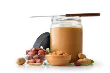 Cam bir kavanozda fıstık ezmesi, metal ve ahşap bir spatula, kabuklu ve beyaz arka planda yapraklı tanecikleri gösteren fıstık ezmesi. Ön görünüm.