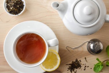 Beyaz seramik bir fincanda limonlu siyah çay, kuru çay yaprakları, metal zerk makinesi ve nane yapraklarıyla dolu tahta bir masada çaydanlık detaylarıyla arka plan. Üst görünüm.