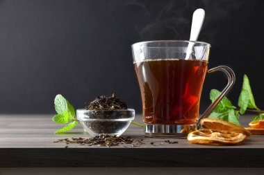 Portakal dilimleri, nane şekeri ve koyu izole edilmiş arka planda bir kase dolusu kuru çay yaprağı olan ahşap bir masanın üzerindeki cam fincanda siyah çay eritimi. Ön görünüm.