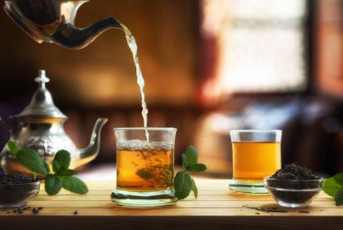 Oturma odasında nane yapraklarıyla süslenmiş bir kase kurutulmuş yaprak çayıyla dolu ahşap bir masanın üzerindeki bir bardağa kadar Fas çayı dökme detayları. Ön görünüm.