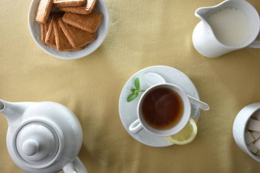 Beyaz seramik bir fincanda çay, çaydanlık, süt sürahisi, şeker küpleri ve bisküviler, bej kumaştan bir masa örtüsü olan beyaz seramik kaplarda. Üst görünüm.