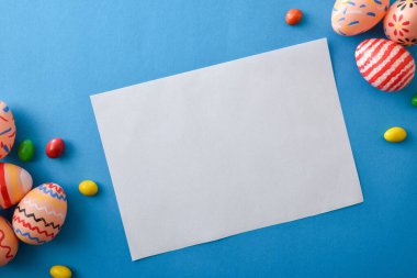 Renkli Paskalya yumurtaları ve parlak mavi arka planda şekerler ile çevrili beyaz bir kartın üst görüntüsü. Bayram tebrikleri ya da davetiyeler için mükemmel.