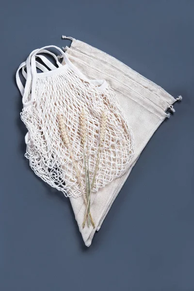 The white reusable string bag and three string sacks for different products on the grey background with three wheat spikelets.