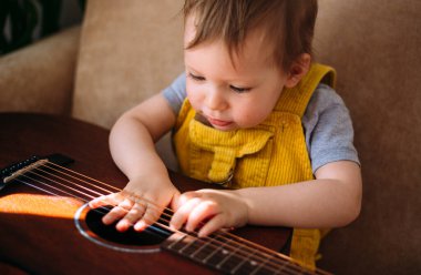 Sevimli bir çocuk evdeki kanepede büyük bir akustik gitarıyla oynuyor.