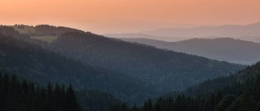 Vadide sisli bir dağ sırasının üzerinde turuncu bir gün batımı. Yamaçlar kozalaklı ormanlarla kaplı, çayır manzaralı..