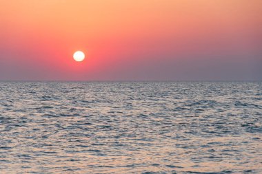 Denizin üzerinde kızıl bir günbatımı, ufukta güneş. Kıyıdan görünen kıyı manzarası.