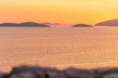 Denizin üzerinde turuncu bir günbatımı. Suda birçok ada var. Tepe manzaralı bir kıyı manzarası. Güneş ufukta batıyor..