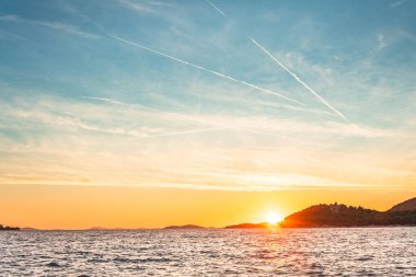 Denizin üzerinde turuncu bir günbatımı. Suda birçok ada var. Kıyıdan görünen bir kıyı manzarası. Güneş ufukta batıyor..