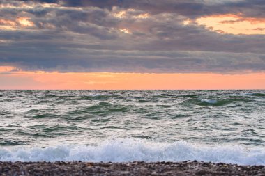 Fırtınalı bir denizin üzerinde portakal rengi gün batımı, ufukta yoğun bulutlar, sahilden gelen deniz dalgalarının görüntüsü akşam geç saatlerde sahilde..