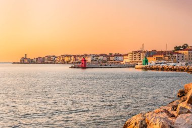 Slovenya 'nın sahil kenti Piran, denizin üzerindeki batan güneşin ışınlarıyla aydınlanıyor. Kıyı manzarası, turuncu bir gökyüzü ile kıyı tarafından görülebilir..