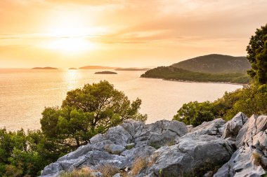 Küçük adalar manzaralı turuncu bir günbatımı. Güneş, kalın bulutların arkasındaki ufkun aşağısında ve kıyı manzarası dağın tepesinden görülebiliyor..