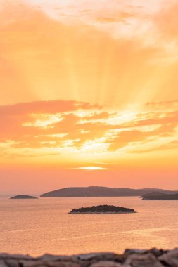 Denizin üzerinde turuncu bir günbatımı. Suda birçok ada var. Tepe manzaralı bir kıyı manzarası. Güneş ufukta batıyor..