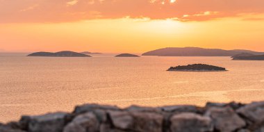Denizin üzerinde turuncu bir günbatımı. Suda birçok ada var. Tepe manzaralı bir kıyı manzarası. Güneş ufukta batıyor..