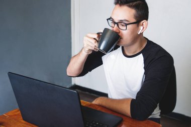 Beyaz adam evde çalışırken siyah bardaktan çay içiyor ve laptopunun önünde oturuyor.