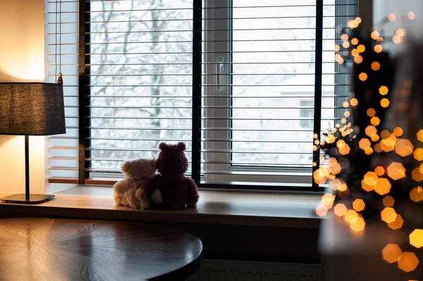 Cozy home evening-two embracing loving teddy bear toys sitting together on wooden windowsill with burning lamp and garland light decor. Living room interior. Filtered horizontal indoor image