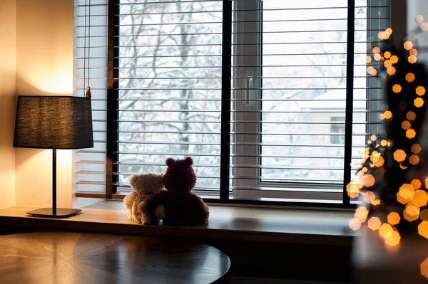 Cozy home evening-two embracing loving  teddy bear toys sitting together on wooden windowsill with burning lamp and garland light decor. Living room interior. Filtered horizontal indoor image