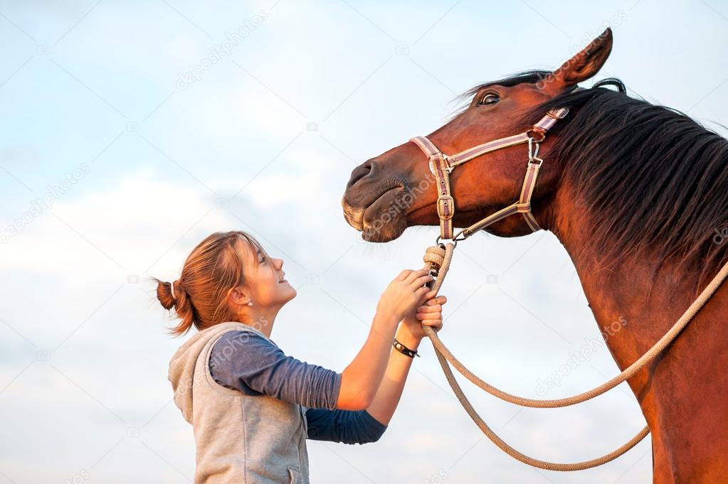 Young cheerful teenage girl calming big brown horse. Outdoors im