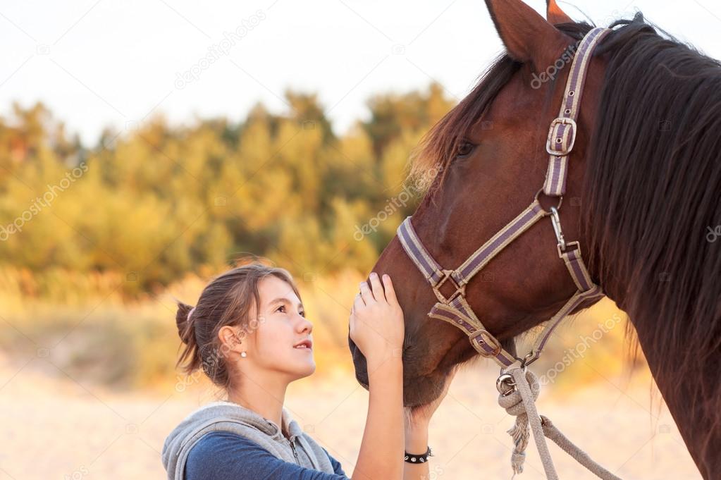 Young cheerful teenage girl stroking brown horse's nose. Outdoor
