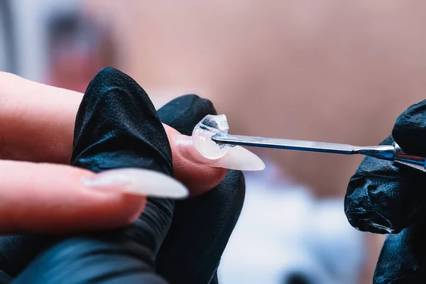Manikür ustası çiviye metal bir spatulayla Poly-gel tabanı uyguluyor. Fırça jeli çivinin tüm yüzeyine dağıtıyor. Yakın çekim, seçici odaklanma