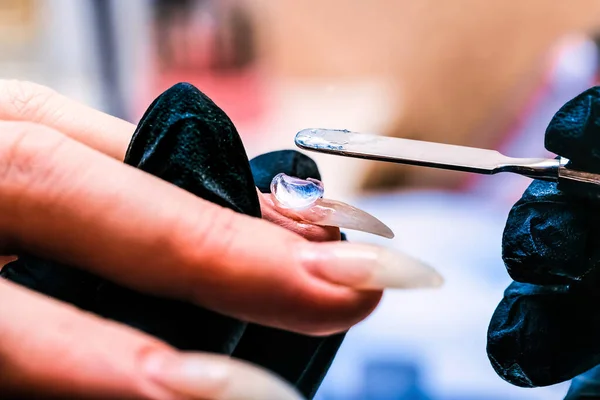 Manikür ustası çiviye metal bir spatulayla Poly-gel tabanı uyguluyor. Fırça jeli çivinin tüm yüzeyine dağıtıyor. Yakın çekim, seçici odaklanma