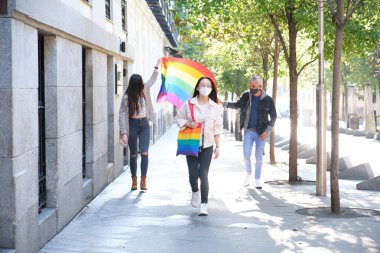 Yüzü maskeli üç arkadaş grubu sokakta LGBT bayrağı sallıyor. Salgın zamanlarında LGBT gurur kutlaması.