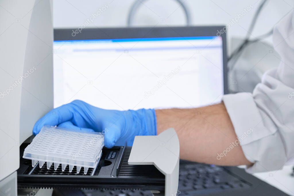 A researcher putting PCR plate on the thermal cycler for DNA amplification. Curve chart on monitor behind. Coronavirus PCR test.
