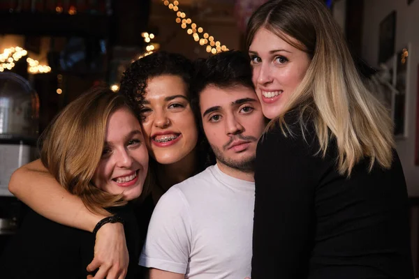 Three smiling young women and one serious young man hugging each others in a bar. Group of friends.