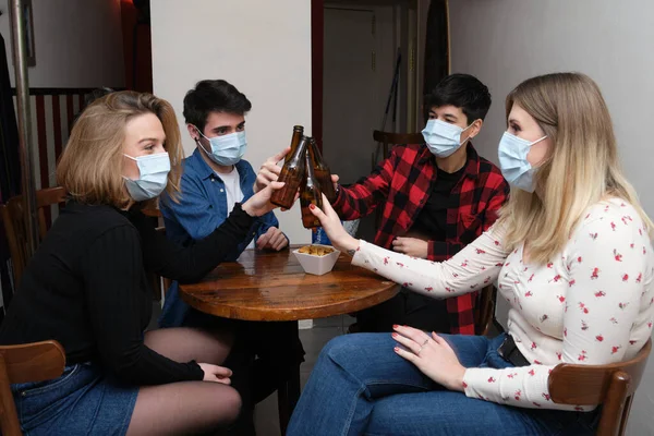 Four friends wearing face masks toasting with beer bottles in a pub. New normal in bars.