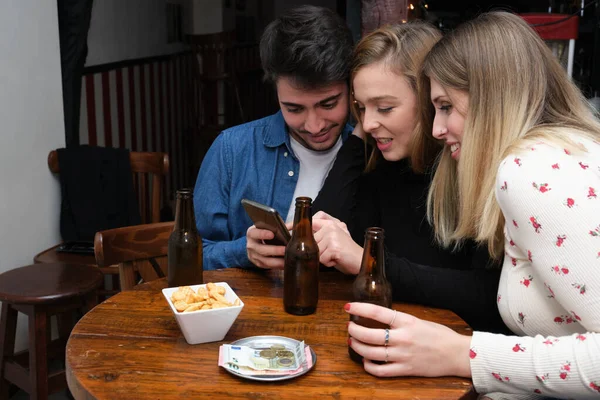三位朋友在智能手机上看到了一些东西 他们的桌上有饮料 零食和钱 酒吧常规概念 — 图库照片
