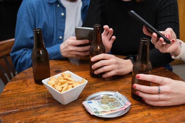 三个不认识的朋友在智能手机上看到了一些东西 他们的桌上有饮料 零食和钱 酒吧常规概念 — 图库照片