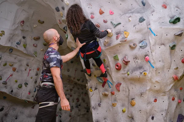 Tırmanma duvarına tırmanırken genç bir kız öğrenciye yardım eden koruyucu yüz maskesi takan bir tırmanış eğitmeni. Yeni normalde öğrenme süreci.