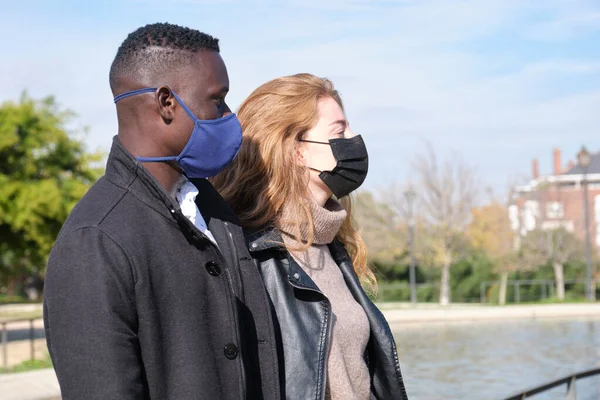 African black man and redhead caucasian woman wearing protective face masks in a park. Young multiracial couple during coronavirus pandemic.